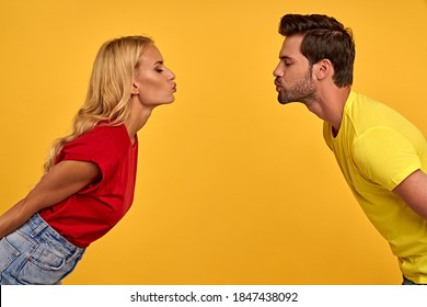 Side Profile View Of Pretty Young Couple Two Friends In Colored T-shirts Isolated On Yellow Wall Background Studio Portrait. Kissing Each Other With Closed Eyes While Standing Face To Face.