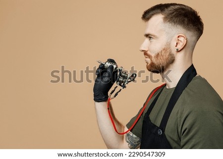 Side profile view tattooer master artist tattooed man wear green t-shirt apron raise up hand hold machine black ink in jar, equipment for making tattoo art on body isolated on plain beige background