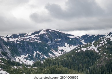 Side profile view of a white floatplane in flight soaring alongside steep, rocky mountainsides patched with snow and alpine greenery.