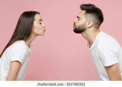 Side profile view young cheerful couple two friends man woman in white basic t-shirts kissing each other with closed eyes while standing face to face isolated on pastel pink background studio portrait