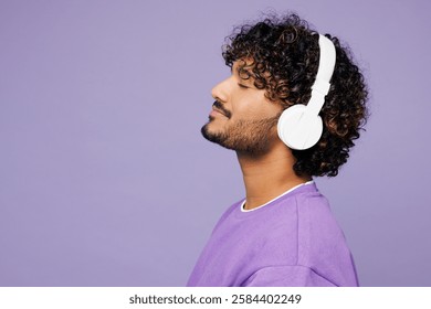 Vista lateral del perfil del joven indio que usa sudadera violeta ropa casual escuchar música en auriculares cerrar los ojos aislados en el retrato de estudio de fondo púrpura claro pastel liso. Concepto de estilo de vida