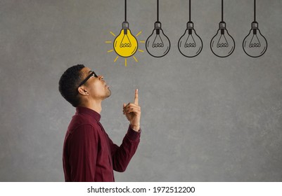 Side profile view young man making choice looking up at several electric light bulbs. Creative Afro American entrepreneur considers different effective business ideas, points finger and picks best one