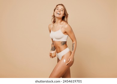 Side profile view young nice lady woman with slim body perfect skin wearing nude top bra lingerie stand apply cream on thigh isolated on plain pastel light beige background. Lifestyle diet fit concept