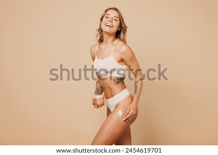 Side profile view young nice lady woman with slim body perfect skin wearing nude top bra lingerie stand apply cream on thigh isolated on plain pastel light beige background. Lifestyle diet fit concept