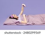 Side profile view young woman wear pyjamas jam sleep eye mask rest relax at home lay down on bed hold use mobile cell phone play games isolated on plain purple background. Good mood night nap concept
