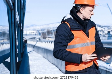 Side profile of worker checking clipboard in snow. Profile view of helmeted man in orange vest holding and reading clipboard on a winter bridge with mountainous backdrop.