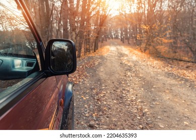 Side rear mirror view of SUV car drive on beautiful dirt gravel forest unpaved road in autumn foliage and bright warm sunlight through woods background. Off road car adventure nature fall journey