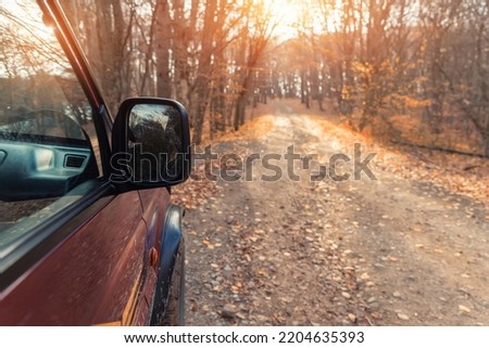 Side rear mirror view of SUV car drive on beautiful dirt gravel forest unpaved road in autumn foliage and bright warm sunlight through woods background. Off road car adventure nature fall journey