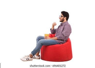 Side Shot Of Handsome Happy Beard Young Man With A Cinema Glasses Sitting On A Red Chair And Eating Popcorn, Guy Wearing Caro Shirt And Jeans, Isolated On White Background