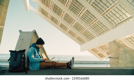 Side Shot Of Modern Urban Millennial Young Man Sit In Public Futuristic Space, Work Remote From Office On Laptop. Startup Employee Work On Idea Project. New Concepts Of Modern Work Environment 