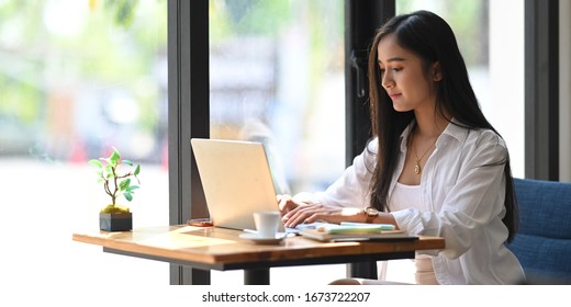 Side shot of young beautiful woman in white shirt typing on computer laptop while sitting at the working wooden table over modern office as background.