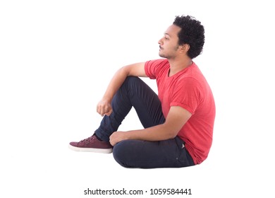 Side shot of a young man wearing a casual outfit and sitting on the floor putting his hands on his knee, isolated on white background