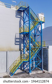 The Side Staircase At The Industrial Building Is Painted Yellow And Blue. Factory Building Outside On A Sunny Day. Outdoor Vertical Photo.