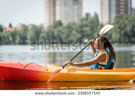 side view of active and charming african american woman in life vest sailing in sportive kayak with paddle on summer weekend day on blurred background