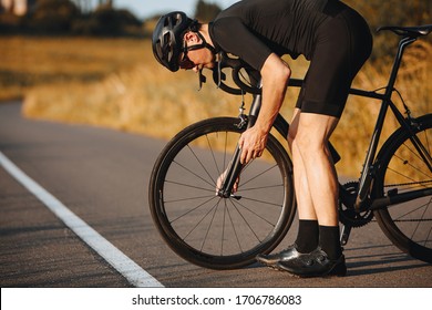 Side View Of Active Man In Sport Clothing Standing Bent Near Black Bike To Fix Wheel. Mature Sportsman In Black Helmet Taking Break For Repairing Vehicle.