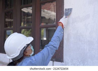 Side View Of An Active Retired Senior Asian Woman Wearing Safety Hard Hat, Goggles, Gloves And Mask, Holding Trowel, Peeling Off Paint Film From Old Wall. DIY Home Renovation  Concept.