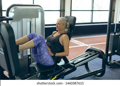 Side View Of Active Senior Caucasian Woman Exercising With Leg Press Machine In Fitness Studio. Bright Modern Gym With Fit Healthy People Working Out And Training