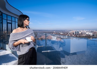 Side view of adorable caucasian woman in sunglasses and stylish wear standing on balcony with cup of coffee in hands and enjoying amazing view on big city. Leisure time and lifestyles concept. - Powered by Shutterstock