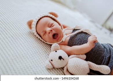 Side View Of Adorable Newborn Baby Wrapped In Wool Scarf And With Little Cap On Head While Lying Next To Toy On Fur Blanket.