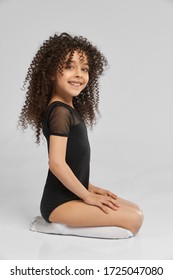 Side view of adorable smiling girl in black sportswear and white knee socks posing while sitting of floor, isolated on gray studio background. Little professional female gymnast with curly hair.