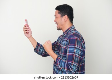 Side View Of Adult Asian Man Clenched Fist Showing Excited Expression When Looking To His Mobile Phone