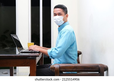 Side View Of Adult Asian Man Wearing Medical Mask Working With Laptop In His Home