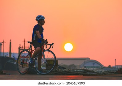 Side View Of Adult Athletic Man On Bike Is Taking Break And Relaxing At Harbor After Long Distance Workout In Sunset Time