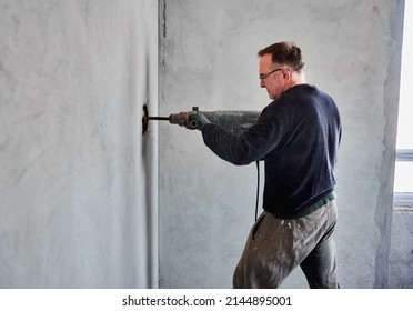 Side View Of Adult Builder With Glasses Who Using Perforator To Make Hole In Gray Wall Inside Room During Overhaul Of Living Space. Reconstruction Work Inside Flat.