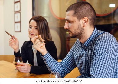 Side View Of Adult Caucasian Man And Woman Couple Sitting By The Table Smoking Cigar And Drinking Cognac Brandy Looking To The Cigarillos In Day Wearing Shirt