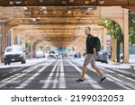 Side view of adult man with backpack walking on pedestrian crosswalk uder elevated railway of public transportation. Chicago, United States
