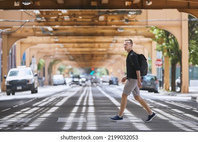 Side view of adult man with backpack walking on pedestrian crosswalk uder elevated railway of public transportation. Chicago, United States
					