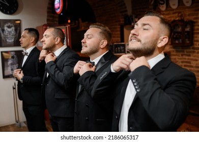 Side view of adult men and bridegroom in black suits, standing and together correcting their bow ties on necks in wedding morning