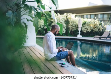 Side View Of Adult Relaxing Man Sitting On Poolside With Feet In Water While Working Remote On Modern Laptop Having Vacation