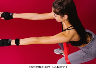 Side view of adult sporty woman with mockup screen of fitness watch in sportswear, exercising with red resistance elastic band squatting on red backgroud.