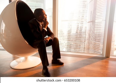 Side view of African american businessman holding hand on chin, sitting in design egg chair enjoying sunset, looking through big office window at city, deep in thoughts, dreaming of new opportunities - Powered by Shutterstock