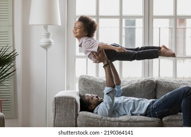 Side View African American Father And Son Playing Active Game, Having Fun, Smiling Dad Lying On Couch, Carrying Little Boy Pretending Flying With Arm Outstretched, Enjoying Leisure Time Together