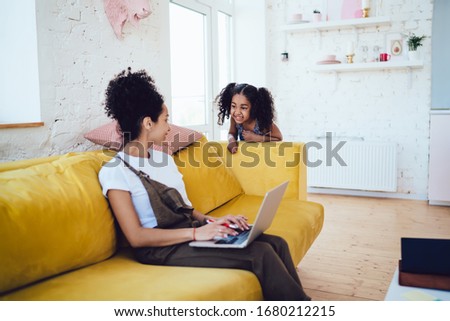 Side view of African American female freelancer in casual clothing with curly hair working on laptop and daughter telling joke