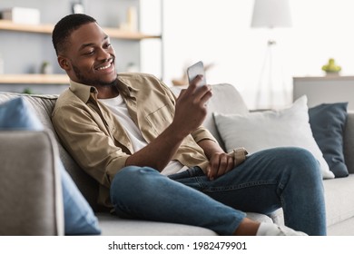 Side View Of African American Guy Using Smartphone Browsing Internet Sitting On Couch At Home. Black Man Texting On Cellphone Or Using New Application On Mobile Phone Indoors