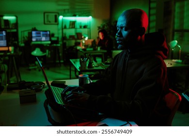 Side view of African American hacker working on laptop with cooling pad in office lit by green lights - Powered by Shutterstock