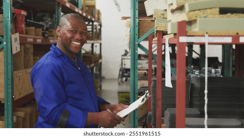 Side view of an African American male worker in a storage warehouse at a factory making wheelchairs, writing on a clipboard, turning, looking to camera and smiling, standing on crutches - Powered by Shutterstock