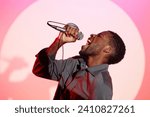 Side view of African American man standing at pastel pink wall of karaoke club and singing passionately into microphone