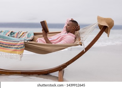 Side View Of African American Man Reading A Book While Relaxing On A Hammock At Beach