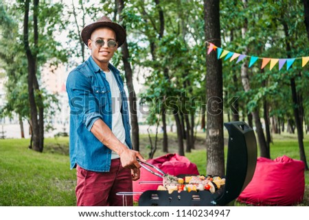 side view of african american man in sunglasses and hat cooking food on grill in park