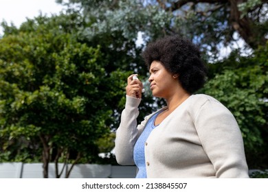 Side View Of African American Mid Adult Woman Using Asthma Inhaler While Standing Against Trees. Unaltered, Healthcare, Asthmatic, Reliever Asthma Inhaler, Inhaling And Illness Concept.