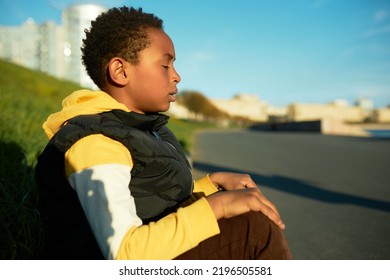 Side View Of African American Peaceful Kid Boy Sitting On Boarder Outdoor At City Street With Closed Eyes, Having Rest, Relaxing, Trying To Calm Down, Breathing Fresh Air On Early Morning