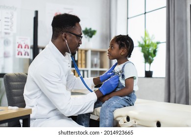 Side view of african american pediatrician using stethoscope for listening little girl's lungs at modern hospital. Qualified doctor doing checkup for female patient.