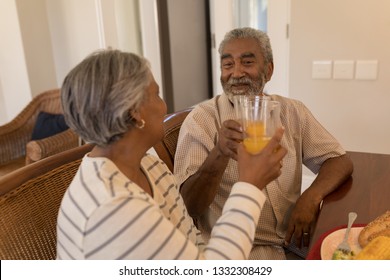 Side View Of African American Senior Couple Toasting Glasses Of Orange Juice While Sitting At The Dining Table At Home