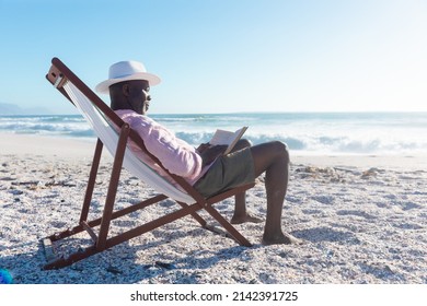 Side view of african american senior man reading book sitting on folding chair at beach. unaltered, hobbies, active lifestyle, enjoyment and holiday concept. - Shutterstock ID 2142391725