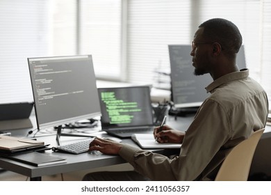 Side view of African American IT specialist typing on computer keyboard and writing in notebook while sitting at desk in cybersecurity office
