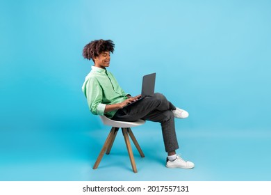 Side View Of African American Student Guy Using Laptop, Surfing Internet While Sitting In Chair On Blue Studio Background, Free Space. E-learning, Online Lectures And Courses